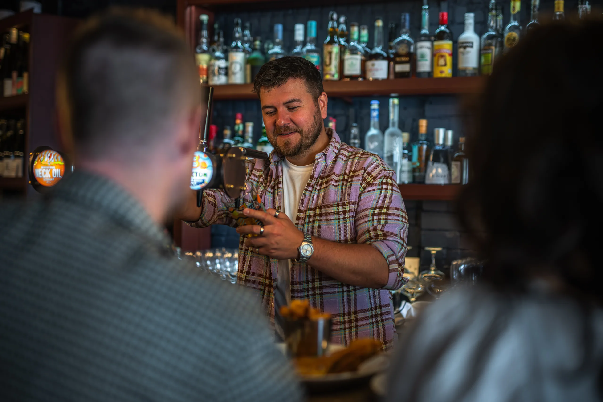 A bartender in a checked shirt pulling a pint at the bar with shelves of spirits behind.