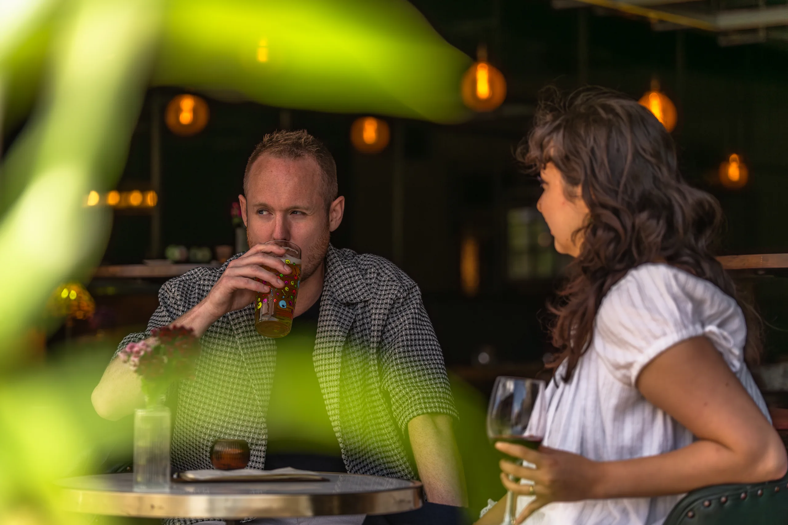 Two guests enjoying drinks at a table, framed by green foliage with warm pendant lighting behind.