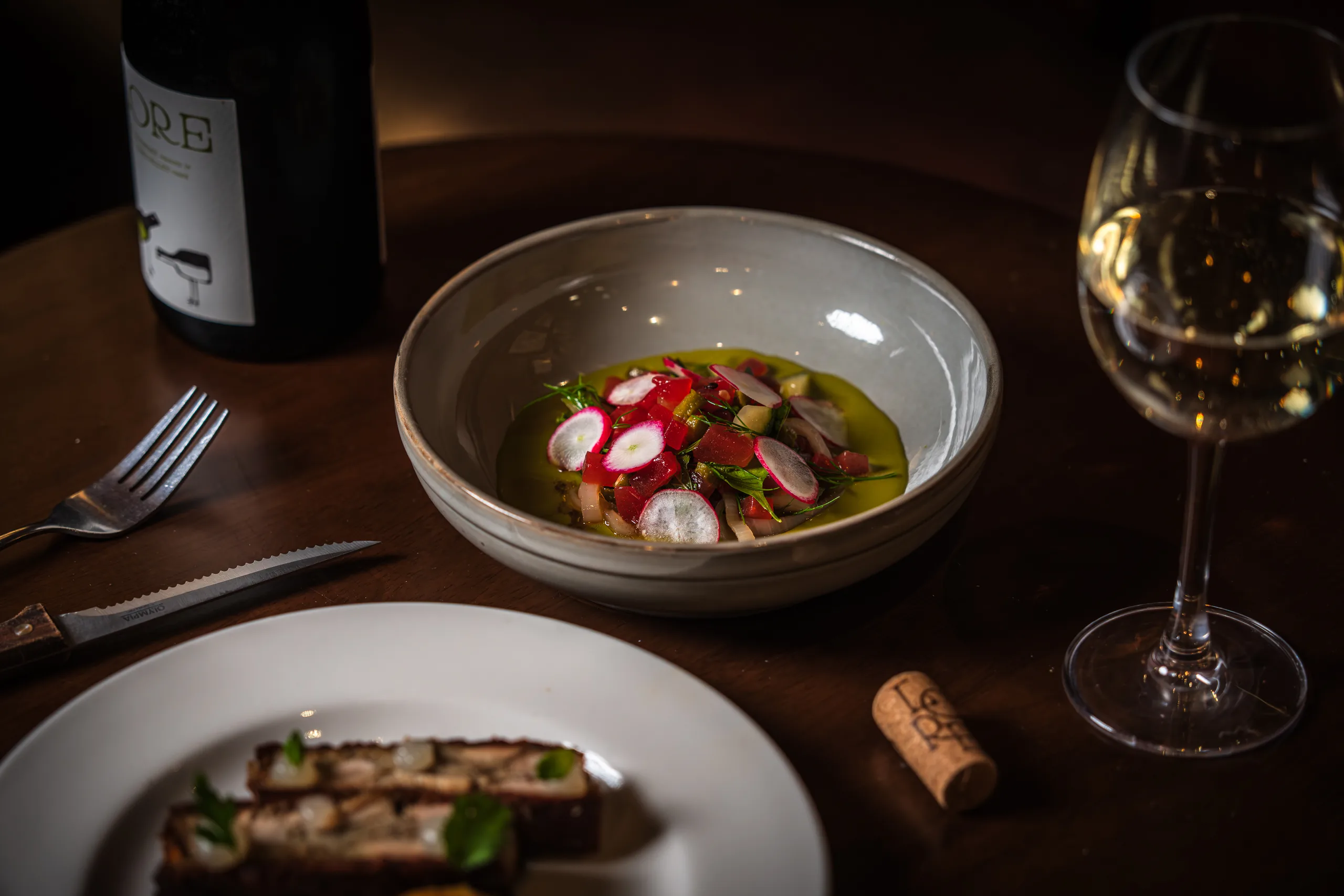 A fresh radish, herb and leaf salad dressed in green oil, with a glass of white wine and a sliced terrine starter nearby.