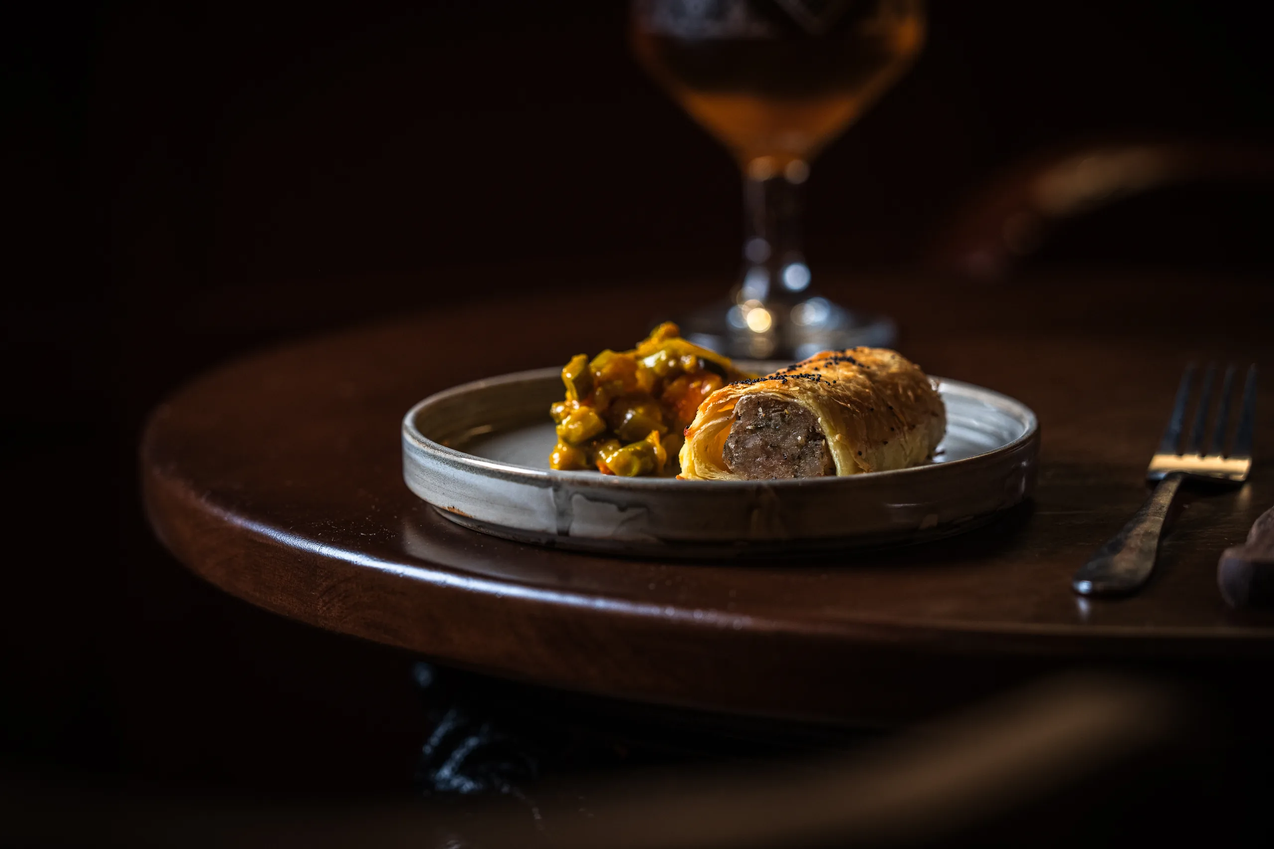 Sausage roll with golden puff pastry sliced to show the filling, served with piccalilli and a glass of ale.