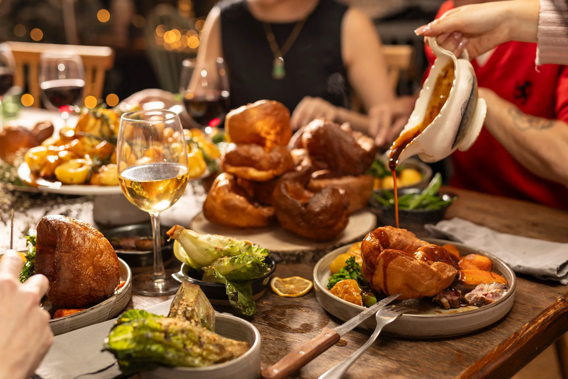 Gravy being poured over a Sunday roast at The Hill, with a stack of Yorkshire puddings, roast potatoes, greens and glasses of wine on a shared table.