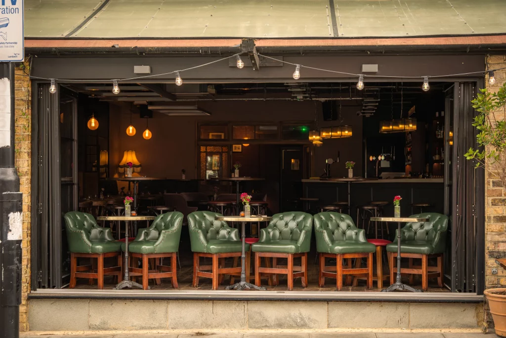 Green leather tub chairs at the open bar window of The Hill with festoon lights and a warm glow at dusk.