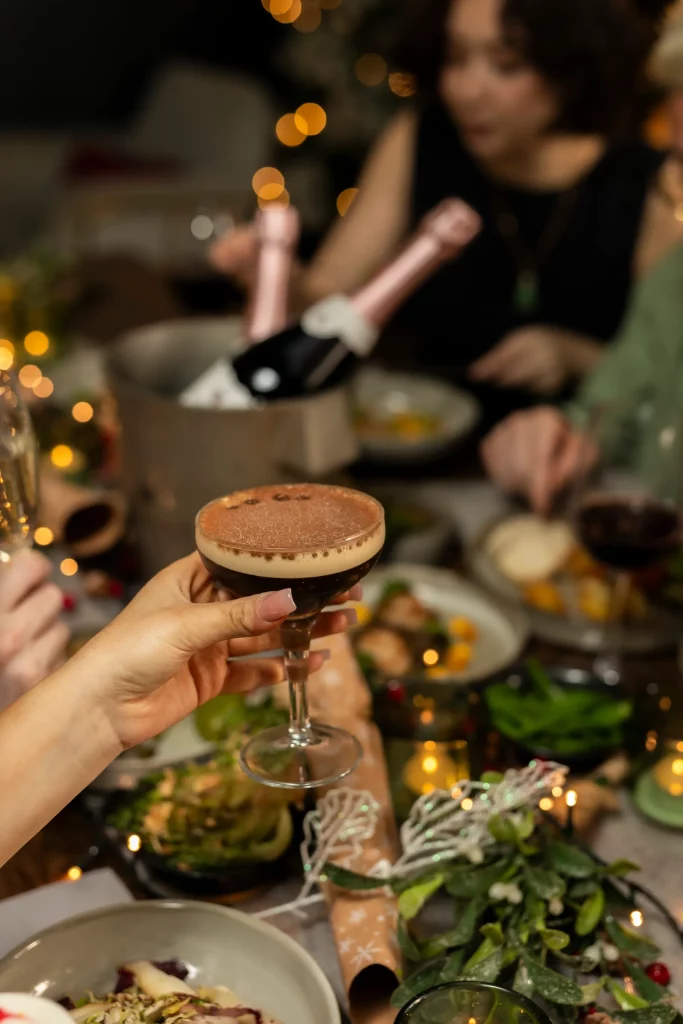 A hand holding up an espresso martini dusted with coffee beans at a festive table, with a bottle of rose prosecco chilling on ice and guests celebrating in the background.